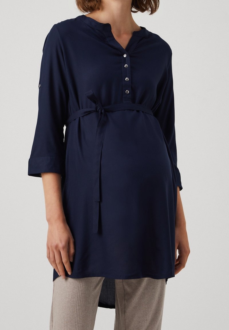 Personne enceinte portant une longue tunique de maternité bleu marine avec un col boutonné et une ceinture à la taille, associée à un pantalon beige clair.