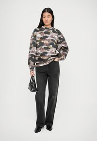 Νεαρή γυναίκα φορώντας ένα πολύχρωμο φούτερ με καμουφλάζ, μαύρα φαρδιά τζιν, μαύρα μυτερά παπούτσια, κρατώντας μία μικρή μαύρη τσάντα.