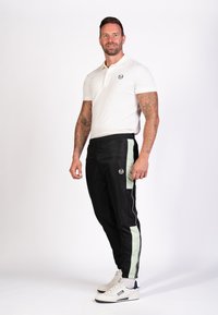 Männeroutfit mit einem taillierten weißen Poloshirt und schwarzen Sporthosen mit mintgrünen Seitenstreifen, kombiniert mit weißen Sneakern.