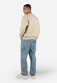 Beige textured sweatshirt with ribbed cuffs and a crew neck, paired with light blue denim jeans featuring a side pocket. White sneakers visible.