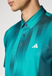 Man som bär en polo med telfärgad och mörkgrön rand, med små perforeringar och en vit Adidas-logotyp på bröstet.