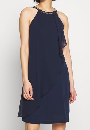 Kvinde iført en ærmeløs mørkeblå kjole med lagdelt chiffon foran og perlebesat halterhals, stående mod en ensfarvet baggrund.