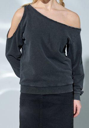 Femme portant un sweat-shirt gris foncé à épaules dénudées avec une découpe sur une manche, associée à une jupe noire sur un fond uni.
