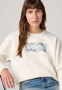Bijela dukserica s plavim cvjetnim Levi's logom na prednjoj strani. Model ima dugu, valovitu kosu i nosi traperice.