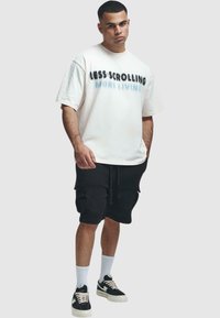 T-shirt blanche surdimensionnée avec le texte noir et bleu "MOINS DE DÉFILEMENT, PLUS DE VIE". Shorts cargo noirs, baskets gris et blanc, chaussettes blanches.