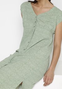 Robe fleurie verte avec des manches courtes, détail de boutons et encolure en V. Texture de tissu douce avec de petits motifs blancs tout au long.