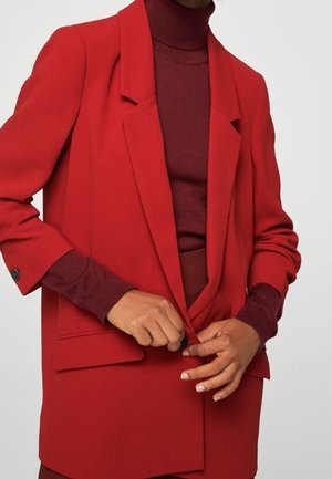 Personne boutonnant un blazer rouge vif porté sur un col roulé rouge foncé, sur un fond uni.