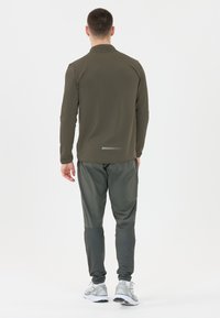 Olivevihreä urheilullinen pitkähihainen paita, jossa on tekstuuri, yhdistetty tummanvihreisiin kapeneviin housuihin. Paidan heijastavat yksityiskohdat, lenkkitossut näkyvissä.