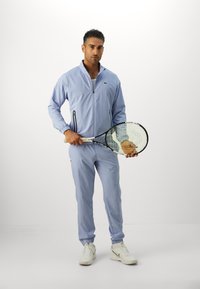 Mand i lyseblå træningstøj holder en tennisracket og en bold, stående foran en ensfarvet hvid baggrund.