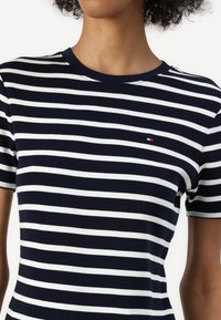 Persoon draagt een navyblauw-wit horizontaal gestreept T-shirt met korte mouwen en een klein logo op de linkerborst.