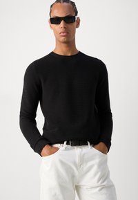 Svart teksturert genser med lange ermer og rund hals, kombinert med hvite bukser og et svart belte. Modellen har på seg rektangulære svarte solbriller.