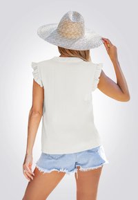 Wit geribbeld topje met cap sleeves en franje-accenten, gecombineerd met versleten denim shorts. Model draagt een brede strohoed.