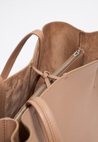 Beige leather handbag with a textured exterior, soft lining, and a zippered pocket. Features flexible handles and a minimalist design.