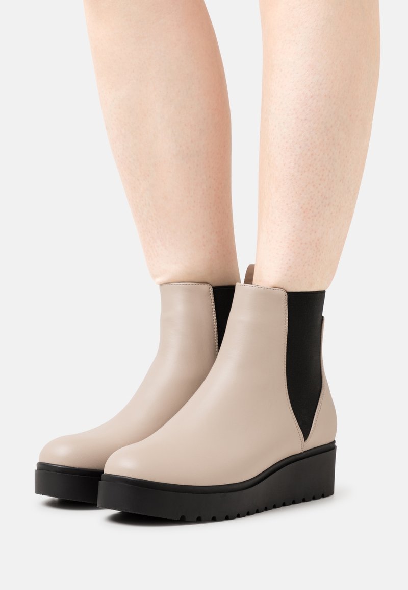 Bottines beiges avec panneaux latéraux élastiques noirs et semelles compensées épaisses noires portées sur des jambes nues sur fond blanc.