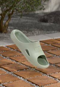 Sandale à enfiler vert clair avec des trous de ventilation, semelle texturée et logo embossé, flottant au-dessus de carreaux en bois et d'un fond en gravier gris.