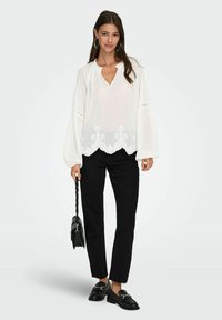 Žena v bílé vyšívané blouse, černých rovný nohavicích, černých mokasínách, s velkými kruhovými náušnicemi a držící černou kabelku.