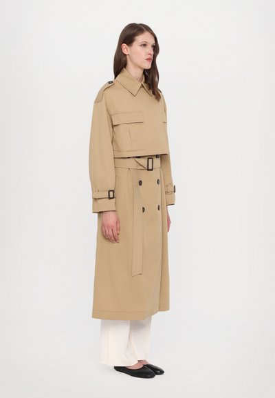 Cappotto trench corto beige con bottoni doppi, spalline e due tasche frontali; abbinato a pantaloni bianchi larghi e scarpe nere.