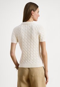 Vit kortärmad stickad tröja med texturerat kabelmönster och ribbad krage, kombinerad med lösa beige linnebyxor.