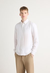 Hvit lin skjorte med knapper, krage, lange ermer og buet kant. Brukt med beige bukser som har en subtil logo detaljer.
