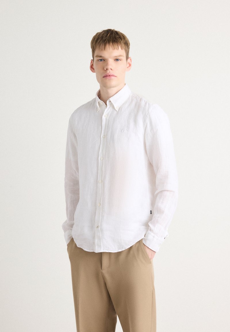 Hvit lin skjorte med knapper, krage, lange ermer og buet kant. Brukt med beige bukser som har en subtil logo detaljer.