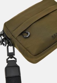 Borsetta in nylon verde oliva con chiusura a zip, dotata di accessori neri e una tracolla texturizzata. Il nome del marchio è impresso sulla superficie.