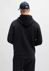 Black hoodie with a snug fit, textured fabric, and a drawstring hood. Worn with a baseball cap, displayed from the back.