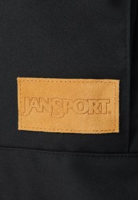 Must mustri seljakott, millel on ristkülikukujuline pruun nahklabel, millel on sisse vajutatud "JANSPORT" tekst, rõhutades vastupidavat materjali ja tekstuuri.