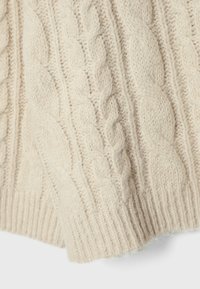 Gros plan d'un pull en laine écru à torsades montrant des motifs tressés et côtelés texturés.