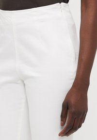 White cotton blend pants with a smooth texture, high-waisted design, and a concealed zipper. Close-up of side and hand detail.