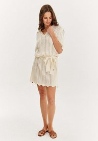 Sukienka dzianinowa w kolorze kremowym, z dekoltem w kształcie litery V, krótkimi rękawami i regulowanym pasem. Posiada wzory przypominające koronkę oraz falbaniany brzeg.