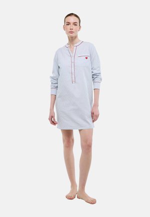 Femme debout pieds nus portant une chemise de nuit rayée bleu clair avec un liseré rouge et un petit cœur rouge sur la poche poitrine.