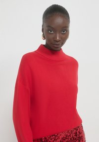 Pull en maille rouge avec un col montant et une coupe ample. Texture lisse, manches longues. Associé à une jupe pailletée dans des teintes de rouge.