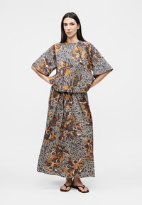 Femme aux longs cheveux foncés portant un haut ample assorti à motif floral et une jupe large bleu marine, orange et blanc, debout, les mains sur les hanches.