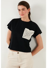 Juodas trumpas rankovės crop top'as su baltu banginiu nėriniu kišenės detalėmis. Minkšta medvilnės medžiaga ir laisvas siluetas. Šviesiai smėliniai kelnes puikiai papildo aprangą.