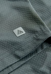 Tissu gris texturé avec un motif à carreaux subtil, comportant une petite étiquette grise avec un logo triangulaire. Matière lisse et légère.