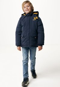 Marinblå isolerad jacka med avtagbar huva, gula detaljer, två framfickor och dragkedjestängning. Stylen är kombinerad med blå jeans och sneakers.