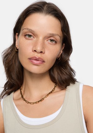Kette aus goldenem Metall mit rechteckigen Gliedern. Das Model trägt ein beiges Tanktop mit einer weißen Unterlage und minimalistischem Schmuck.