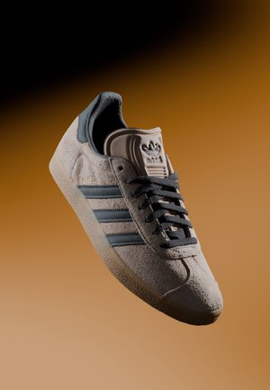 Beżowy sneaker Adidas Gazelle z czarnymi sznurowadłami i paskami, chropowatą zamszową cholewką oraz gumową podeszwą na gradientowym brązowym tle.