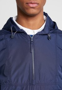 Marinblå huva jacka med dragkedja fram, justerbar dragsko och en lätt, slät väv. Utsmyckad med en kontrasterande dragkedja.