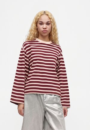 Jeune personne aux longs cheveux blonds bouclés porte un t-shirt ample à manches longues rayé bordeaux et blanc et un pantalon argenté brillant sur fond blanc.