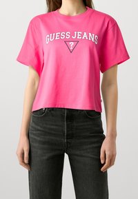 Růžové bavlněné zkrácené tričko s krátkými rukávy, s bílým nápisem "GUESS JEANS" a trojúhelníkovým logem. Spojeno s tmavými džíny.