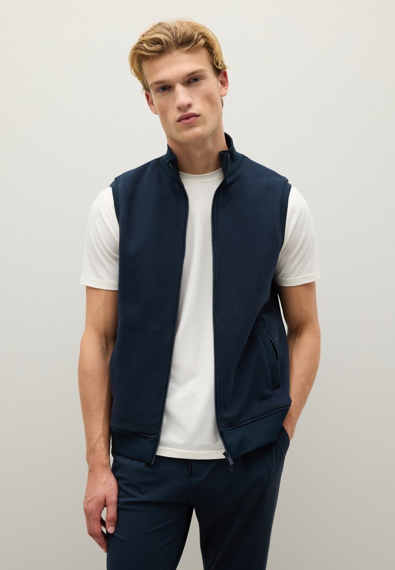 Gilet zip-up blu navy realizzato in tessuto morbido, con colletto alto, due tasche laterali e una cerniera liscia. Indossato sopra una maglietta bianca.