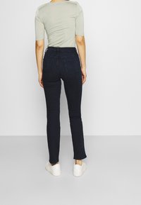 Jeans ajustados de color azul oscuro, con bolsillos traseros, cintura alta y una textura de denim suave, combinados con zapatillas blancas y una blusa verde claro.