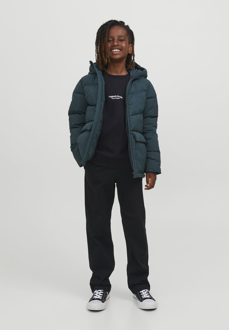Giacca a vento verde scuro con cappuccio, chiusura con zip e design trapuntato. Indossata sopra un felpa blu navy e pantaloni neri, abbinata a sneakers nere.