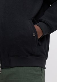 Sweatshirt preto com fecho, feito de um tecido texturizado, com punhos canelados e um bolso lateral segurado por um botão. Usado com calças verdes.