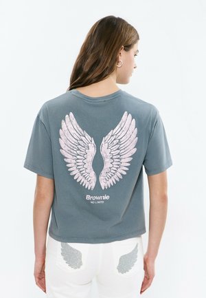 Young woman with long brown hair wears a gray t-shirt featuring large white wings and "Brownie NO LIMITS" text on the back. White pants have wing designs.