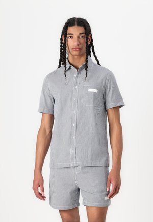 Stribet kortærmet skjorte med knapper og matchende shorts; farverne er marineblå og hvid. Har en brystlomme og diskret branding.