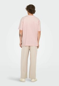 Man with curly hair, wearing a light pink oversized T-shirt, beige wide-leg pants, and white sneakers, standing facing away on white background.