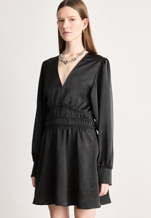 Robe noire avec un décolleté en V, de longues manches bouffantes et une taille smockée. Elle présente un tissu texturé et une jupe fluide, complétée par des accessoires en argent.