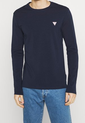 Homme portant une chemise manches longues bleu marine avec un petit logo triangulaire sur la poitrine, associée à un jean bleu clair, debout devant un fond blanc.